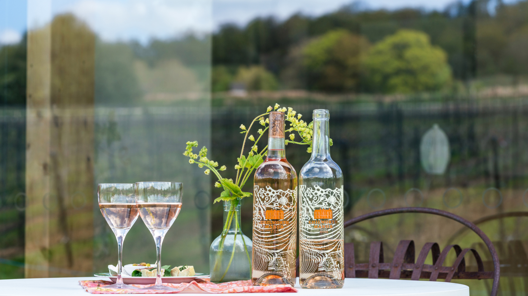 two glasses of still 2025 English rosé wine filled on a white garden table next to two Chilworth Manor Vineyard vintage rosé bottles and a fresh flower vase, overlooking a leafy vineyard.