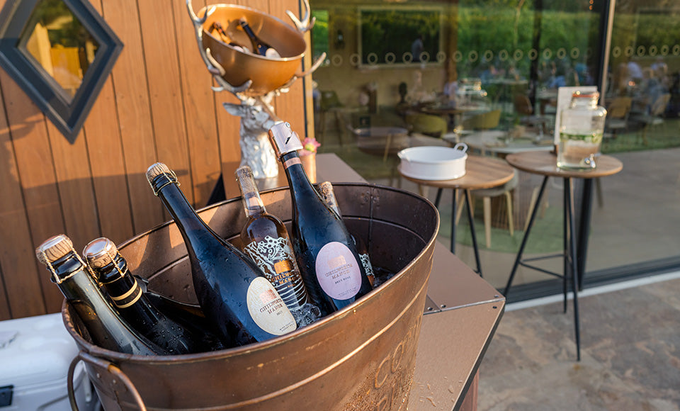 Chilled bottles of Vintage Chilworth Manor English sparkling white and rose wine, with corked bottles of still rosé resting in a copper ice bucket outside the watch house tasting barn, set up for a vineyard event in the Surrey Hills with the display stags head showcasing more bottles.